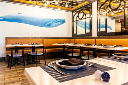 An inviting nautical-themed restaurant dining area featuring a large blue whale mural on a white wall. The seating includes a continuous L-shaped tan leather banquette and dark wood chairs with blue and white patterned cushions. White tables are set with blue striped placemats, dark napkins, and shell-shaped salt and pepper shakers. The space is accented by industrial-style Edison bulb lighting and arched black-framed mirrors that reflect the whale mural.